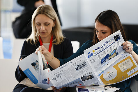 Dos mujeres sentadas leyendo un número del periódico «interpack news», centradas en los contenidos relacionados con el evento.